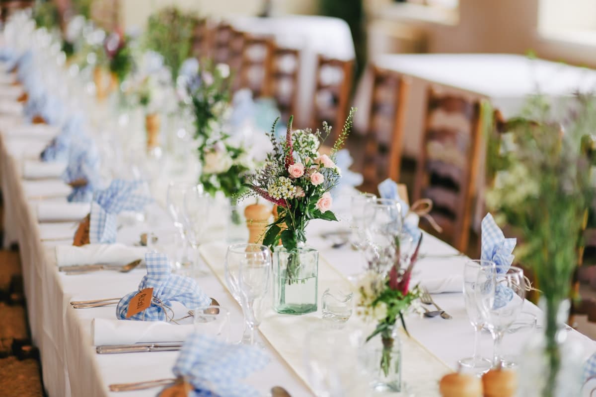 A long reception table set with pale roses and greenery in glass vases and blue gingham napkins